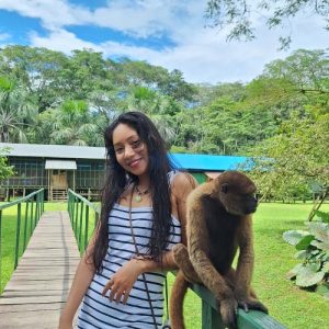 Turista sonriente con un Mono Choro (Lagothrix lagothricha) posado a su lado sobre la baranda de un puente de acceso a un lodge rústico, promocionando un tour de dos días en Iquitos.