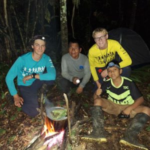Grupo de personas reunidas alrededor de una fogata en la selva amazónica durante la noche, con tienda de campaña al fondo.