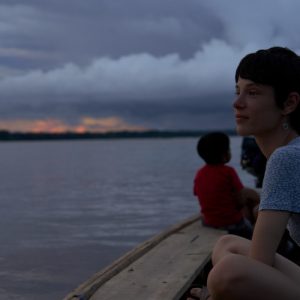 Dos personas navegan en bote por un río amazónico durante el atardecer, rodeados de cielo nublado y vegetación tropical.
