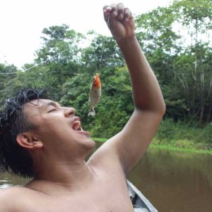 Joven sosteniendo una piraña recién pescada en la Amazonía, simulando comerla, rodeado por el río y la selva.