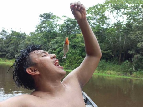 Joven sosteniendo una piraña recién pescada en la Amazonía, simulando comerla, rodeado por el río y la selva.