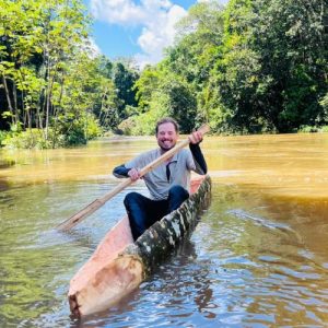 Persona navegando en una canoa artesanal por un río amazónico, rodeado de vegetación tropical y cielo despejado.