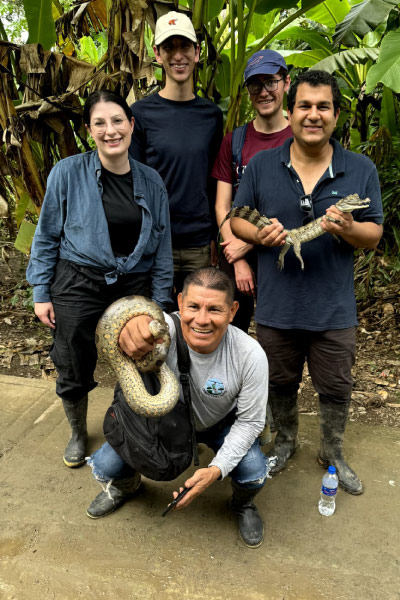 Grupo de investigadores y visitantes sostiene un caimán y una serpiente durante una expedición en la selva amazónica, rodeados de vegetación tropical.