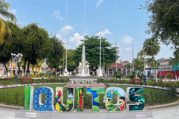 Bienvenidos a Iquitos: La Puerta de Entrada a la Selva Amazónica Letras turísticas coloridas de "IQUITOS" en la Plaza de Armas de la ciudad, con el obelisco y palmeras bajo un cielo despejado