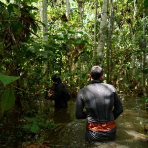 Dos exploradores con equipo técnico avanzado atravesando a pie una zona de selva inundada con el agua a la cintura, rodeados de densa vegetación tropical en Iquitos.