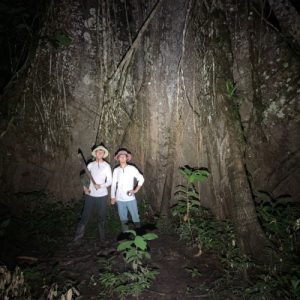 Dos exploradores con linternas y machete posando frente a la base de una ceiba gigante de raíces tabulares durante una caminata nocturna en la Reserva Nacional Pacaya Samiria.